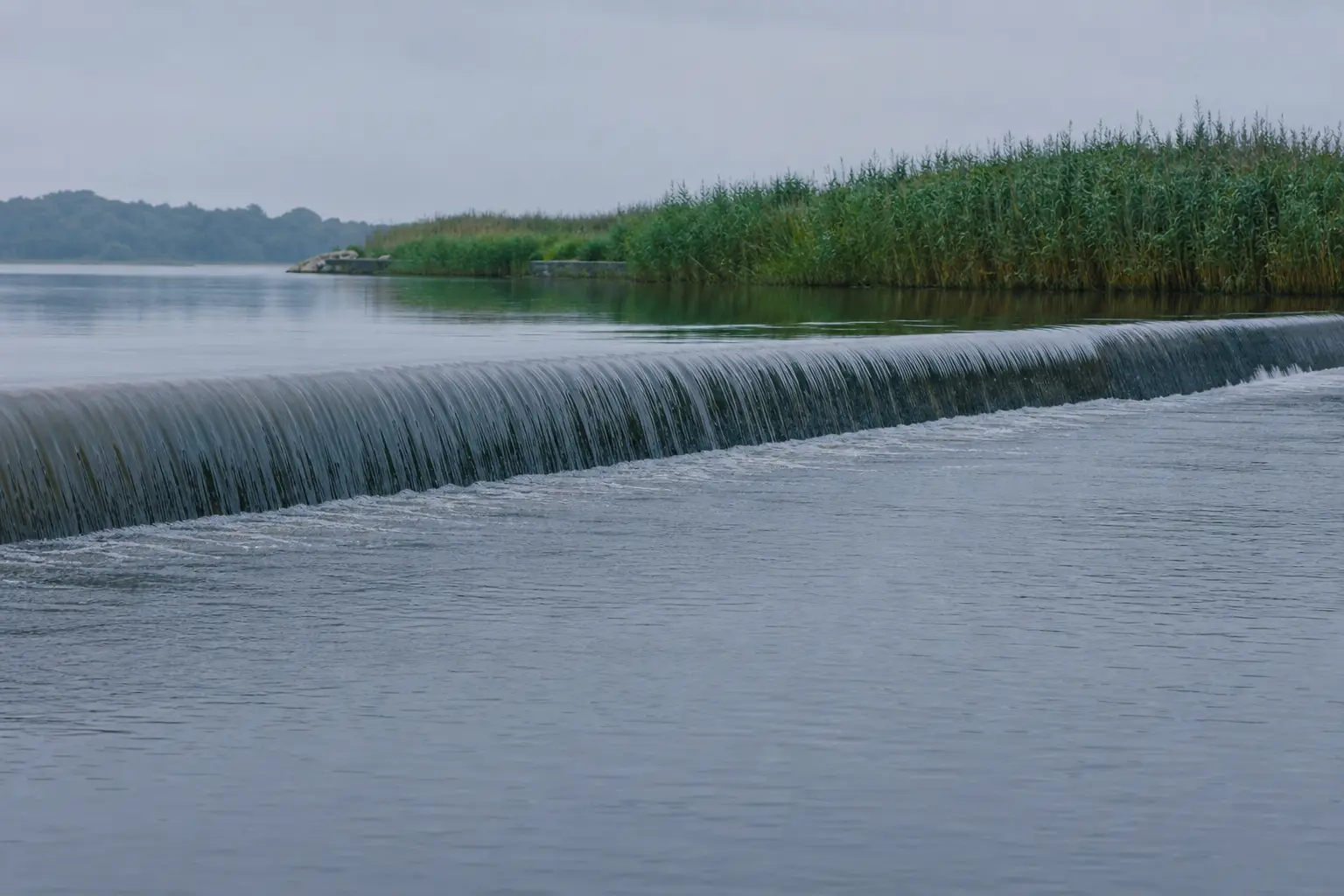 A concrete weir with water sliding over its lip under grey light